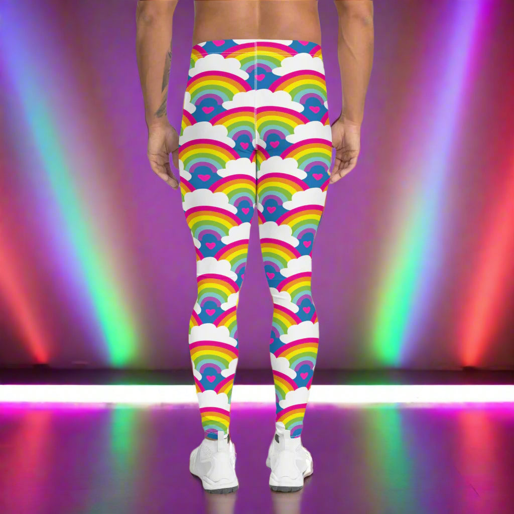 Man from the waist down wearing men's Rainbow Love funky festival yoga leggings standing facing away with his hands by his sides. The man is standing on a dance floor with colourful stage light either side of him in red and green which are reflected on the floor. The background is hazy mixed with glow from the coloured lights. The rainbow love pattern features repeating rainbows and white fluffy clouds with a small pink heart beneath each rainbow. The background colour is royal sky blue