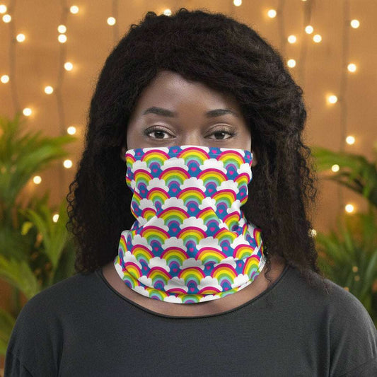 Woman with afro hair facing to the front wearing a grey top and a vibrant rainbow love neck gaiter around her neck, covering her mouth and nose. In the background are some fairy lights and plants. The rainbow love pattern features repeating rainbows and white fluffy clouds with a small pink heart beneath each rainbow. The background colour is royal sky blue.