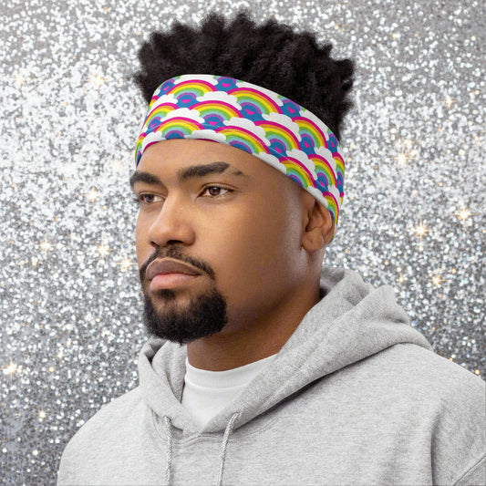 Man with short afro hair facing to the left wearing a grey top and a vibrant rainbow love neck gaiter around his head as a headband. In the background are some fairy lights and plants. The rainbow love pattern features repeating rainbows and white fluffy clouds with a small pink heart beneath each rainbow. The background colour is royal sky blue.
