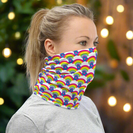 Woman with blonde hair facing to the right wearing a grey top and a vibrant rainbow love neck gaiter around her neck, covering her mouth and nose. In the background are some fairy lights and plants. The rainbow love pattern features repeating rainbows and white fluffy clouds with a small pink heart beneath each rainbow. The background colour is royal sky blue.