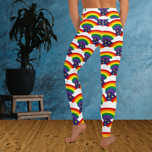 Woman from the waist down wearing high-waist Rainbow Sparkle Pride funky festival yoga leggings standing facing backwards with her hands in front of her. The woman is standing on a wooden floor in front of a textured blue background with a leafy plant in a black pot on a small plant stand to the left of the image. The rainbow sparkle pattern features repeating rainbows and white fluffy clouds with three small yellow stars beneath each rainbow. The background colour is navy blue