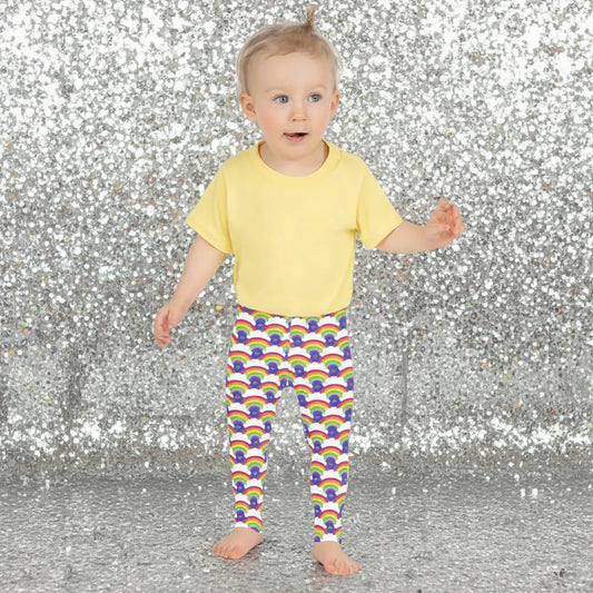 A toddler wearing kids size royal blue rainbow love funky festival leggings and a pale yellow t-shirt, stands facing forwards in front of a calm lake, surrounded by green trees which reflect across the lake. A rainbow arcs above her in the sky. The rainbow love pattern features repeating rainbows and white fluffy clouds with a small pink heart beneath each rainbow. The background colour is royal sky blue.