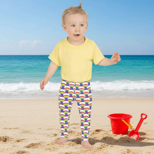 A toddler wearing kids size royal blue rainbow love funky festival leggings and a pale yellow t-shirt, stands facing forwards in front of a calm lake, surrounded by green trees which reflect across the lake. A rainbow arcs above her in the sky. The rainbow love pattern features repeating rainbows and white fluffy clouds with a small pink heart beneath each rainbow. The background colour is royal sky blue.