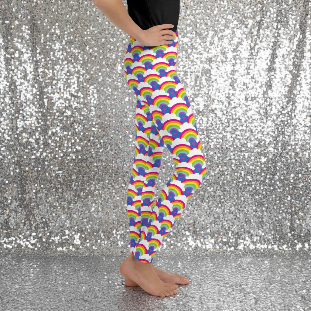 A young person from the waist down wearing youth size royal blue rainbow love funky festival leggings, stands facing to the right. They are standing in front of a calm lake, surrounded by green trees which reflect across the lake. The rainbow love pattern features repeating rainbows and white fluffy clouds with a small pink heart beneath each rainbow. The background colour is royal sky blue.