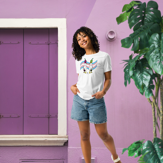 Woman posing casually facing forwards, wearing a fitted white Sassy Seagull Trans Pride Flag LGBTQ+ Pride T-Shirt and stonewash blue jean shorts. She is standing against a purple and white background with a tall green leafy plant to the front right. The t-shirt is white with short sleeves. The graphic shows two cartoon style seagulls wearing colourful hats walking away in front of a trans pride flag. It is a soft playful design for everyday wear