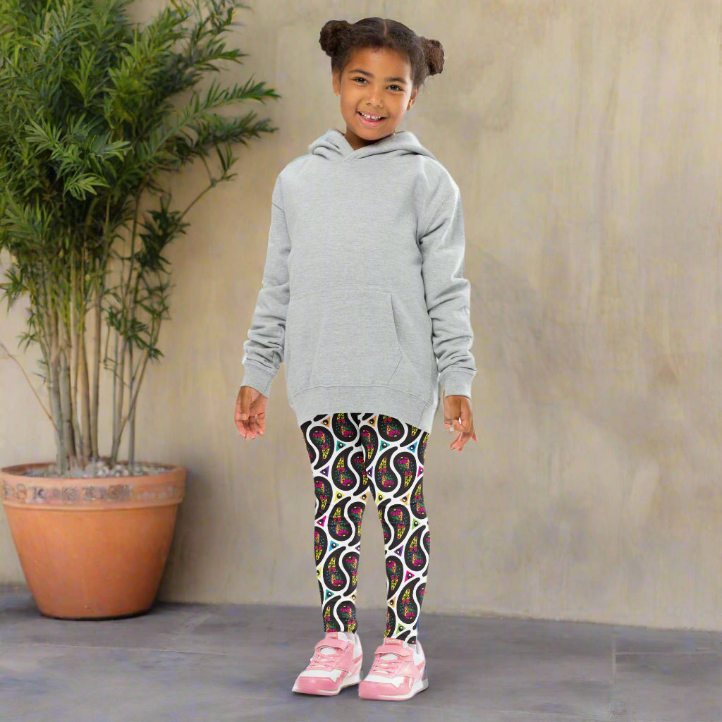 A young girl poses facing forwards, outside in a mediterranean looking setting. She is smiling and standing next to a tall plant in a terracotta pot. She is wearing white paisley party funky festival leggings, and a pale yellow t-shirt. The paisley party pattern is a colourful repeating large paisley pattern, made up of vibrant colours including pink, orange, yellow, green, blue and purple. In between the paisley print are colourful triangles in the same colours. The pattern background is white.