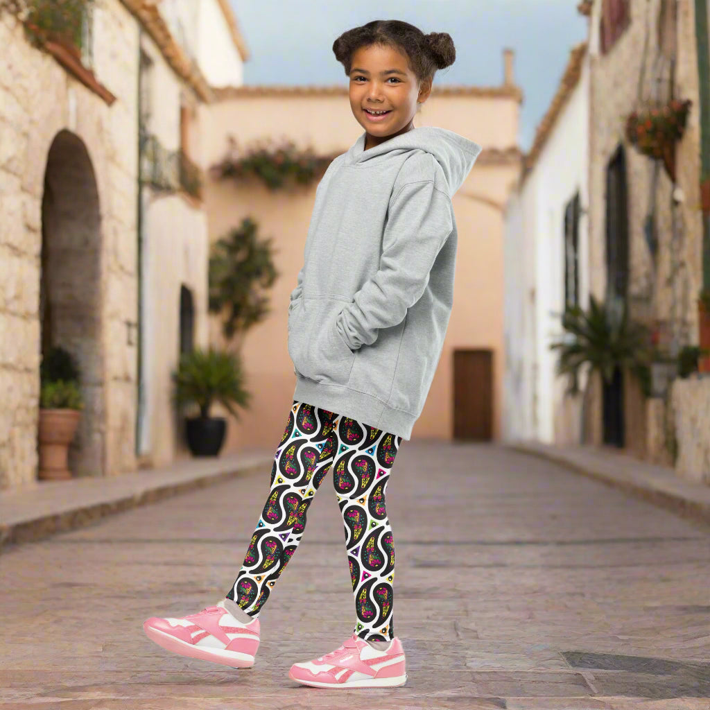 A young girl poses facing to the left outside in a mediterranean looking setting. She is smiling and standing in a quaint narrow street. She is wearing white paisley party funky festival leggings, and a pale yellow t-shirt. The paisley party pattern is a colourful repeating large paisley pattern, made up of vibrant colours including pink, orange, yellow, green, blue and purple. In between the paisley print are colourful triangles in the same colours. The pattern background is white.