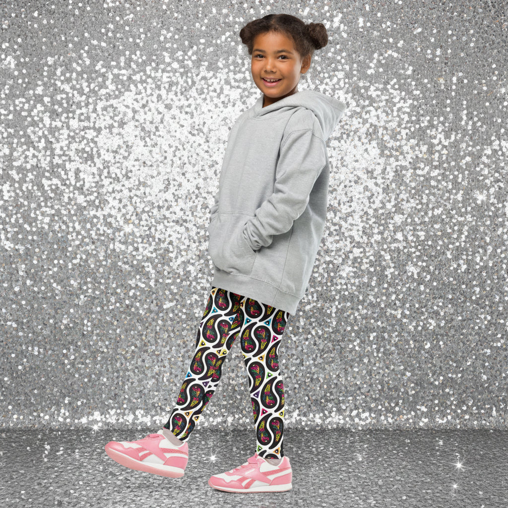 A young girl poses facing to the left outside in a mediterranean looking setting. She is smiling and standing in a quaint narrow street. She is wearing white paisley party funky festival leggings, and a pale yellow t-shirt. The paisley party pattern is a colourful repeating large paisley pattern, made up of vibrant colours including pink, orange, yellow, green, blue and purple. In between the paisley print are colourful triangles in the same colours. The pattern background is white.