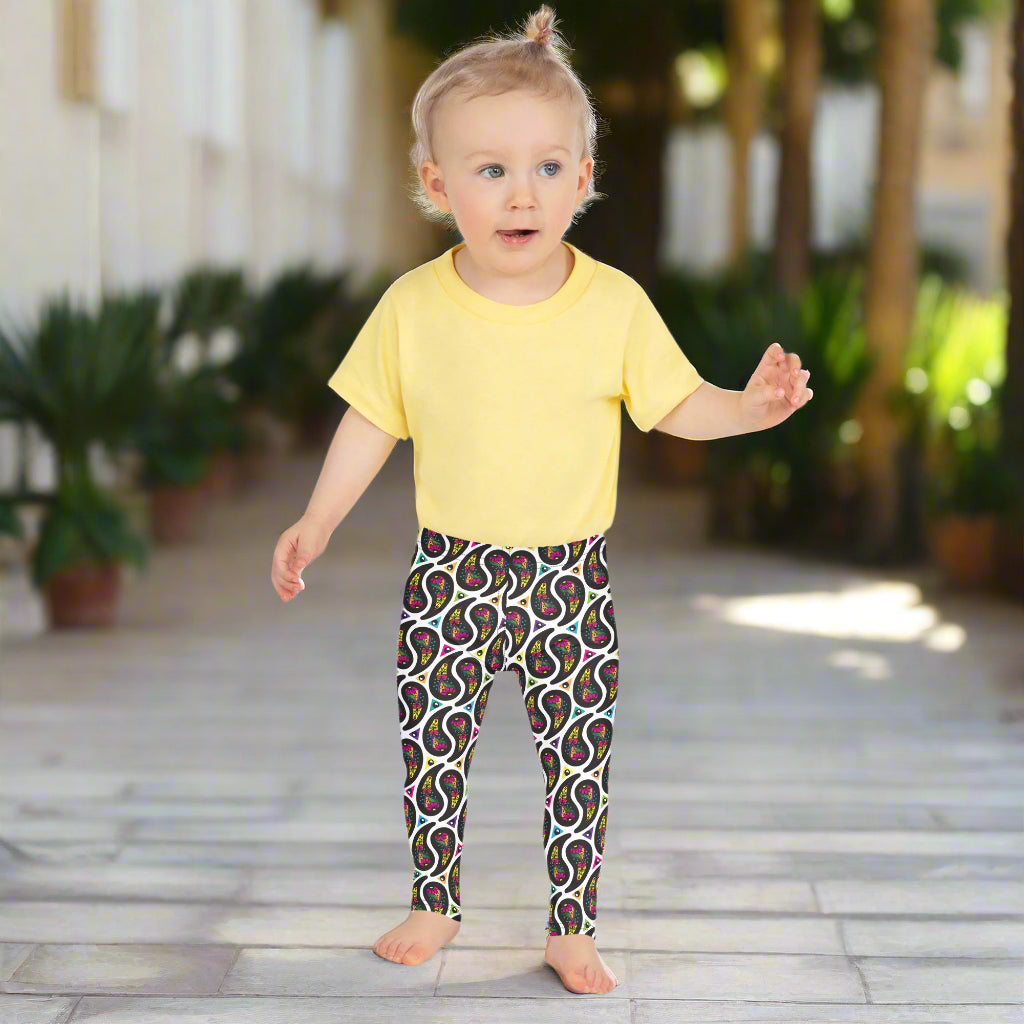 A toddler poses facing forwards outside in a mediterranean looking setting. There are lots of leafy palm type plants in the background. She is wearing white paisley party funky festival leggings, and a pale yellow t-shirt. The paisley party pattern is a colourful repeating large paisley pattern, made up of vibrant colours including pink, orange, yellow, green, blue and purple. In between the paisley print are colourful triangles in the same colours. The pattern background is white.