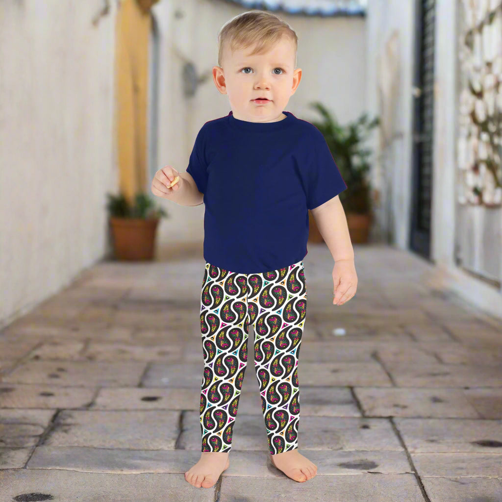 A toddler poses facing forwards outside in a mediterranean looking paved garden area. There are leafy plants in the background. He is wearing white paisley party funky festival leggings, and a pale yellow t-shirt. The paisley party pattern is a colourful repeating large paisley pattern, made up of vibrant colours including pink, orange, yellow, green, blue and purple. In between the paisley print are colourful triangles in the same colours. The pattern background is white.