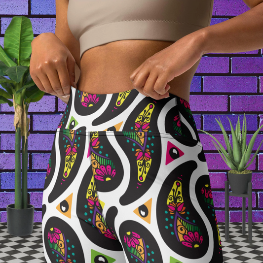 Close up of a woman from the chest down to her thighs wearing white Paisley Party high-waist funky festival yoga leggings standing facing slightly to the left with her hands by her waist. The woman is standing on a black and white tiled floor in front of a vivid purple brick wall with 2 leafy green plants on the floor, one on either side.The Paisley Party pattern is a repeating paisley pattern. The paisley shapes are filled with decorative detail in a variety of bright colours against a white background.