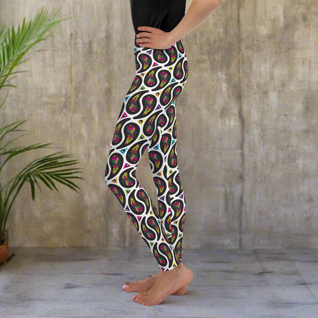 A young person poses facing to the left with one hand on their hip. They are standing in an interior space which looks mediterranean-esque. There are leafy plants. They are wearing white paisley party funky festival leggings, and a black t-shirt. The paisley party pattern is a colourful repeating large paisley pattern, made up of vibrant colours including pink, orange, yellow, green, blue and purple. In between the paisley print are colourful triangles in the same colours. The pattern background is white.
