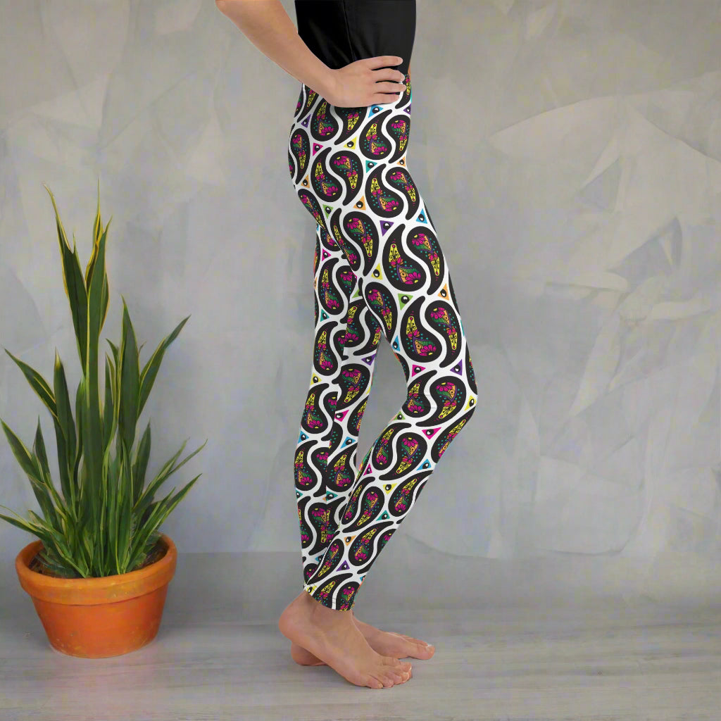 A young person poses facing to the right with one hand on their hip. They are standing in an interior space. There is a leafy plant in a terracotta pot. They are wearing white paisley party funky festival leggings, and a black t-shirt. The paisley party pattern is a colourful repeating large paisley pattern, made up of vibrant colours including pink, orange, yellow, green, blue and purple. In between the paisley print are colourful triangles in the same colours. The pattern background is white.