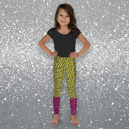 A young girl wearing kids size yellow and pink leopard glamour funky festival leggings and a black t-shirt stands facing forwards. She is smiling and standing in front of a black and white city street scape. This design features an animal print pattern in black, grey and white, printed at a medium scale. The top 3/4 background is bright yellow with the bottom 1/4 bright pink
