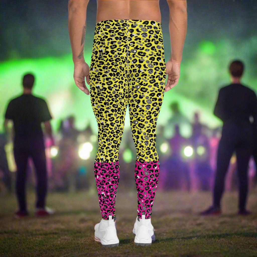 Man from the waist down wearing mens Yellow and Pink Leopard Glamour funky festival yoga leggings standing facing away with his hands by his sides. The man is wearing white trainers and standing outside at a festival after dark with other festival goers and lights in the background. This design features an animal print pattern in black, grey and white, printed at a medium scale. The top 3/4 background is bright yellow with the bottom 1/4 bright pink