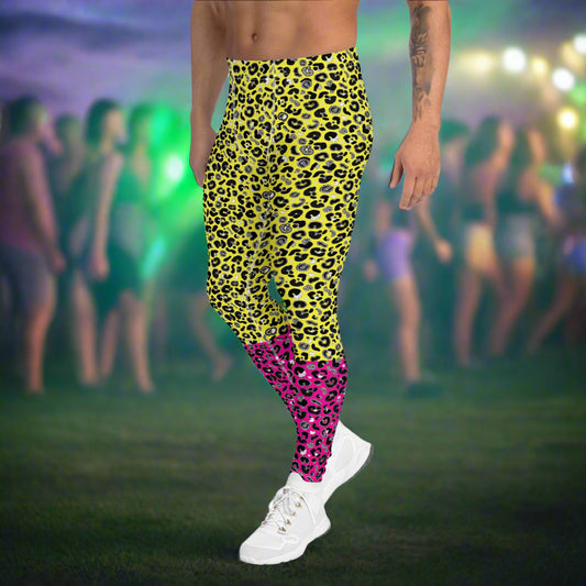 Man from the waist down wearing mens Yellow and Pink Leopard Glamour funky festival yoga leggings standing facing to the left with his hands by his sides. The man is wearing white trainers and standing outside at a festival after dark with other festival goers and lights in the background. This design features an animal print pattern in black, grey and white, printed at a medium scale. The top 3/4 background is bright yellow with the bottom 1/4 bright pink