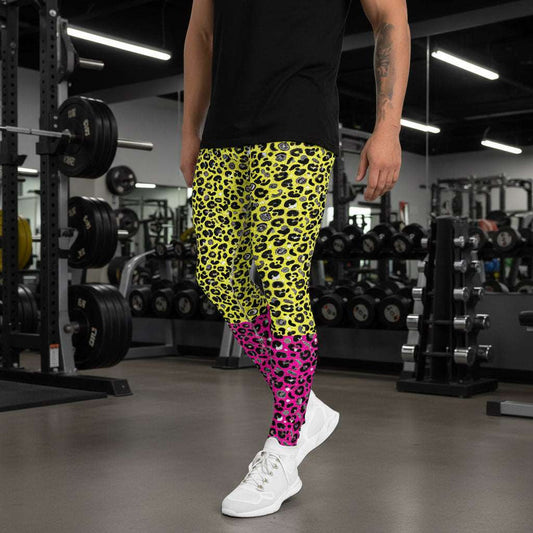 Man from the waist down wearing mens Yellow and Pink Leopard Glamour funky festival yoga leggings standing facing to the left with his hands by his sides. The man is wearing white trainers and standing outside at a festival after dark with other festival goers and lights in the background. This design features an animal print pattern in black, grey and white, printed at a medium scale. The top 3/4 background is bright yellow with the bottom 1/4 bright pink