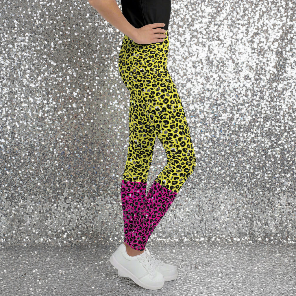 A young person from the waist down wearing youth size yellow and pink leopard glamour funky festival leggings stands facing to the right. They are wearing white trainers and have one hand on their hip. They are standing in front of a black and white city street scape. This design features an animal print pattern in black, grey and white, printed at a medium scale. The top 3/4 background is bright yellow with the bottom 1/4 bright pink