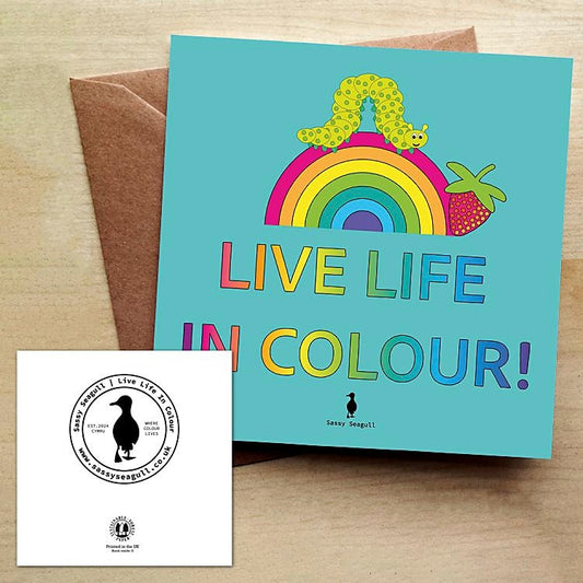 Colourful greeting card with a rainbow, a caterpillar and a strawberry, with the text "Live Life in Colour" on a blue background. The card is on a brown envelope on a wooden table.