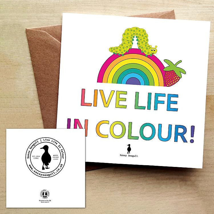 Colourful greeting card with a rainbow, a caterpillar and a strawberry, with the text "Live Life in Colour" on a white background. The card is on a brown envelope on a wooden table.