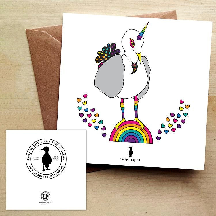 White greeting card with a rainbow unicorn seagull standing on a rainbow with rainbow coloured hearts. Card is on a brown envelope on a brown table.