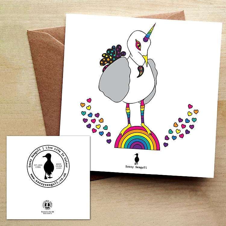 White greeting card with a rainbow unicorn seagull standing on a rainbow with rainbow coloured hearts. Card is on a brown envelope on a brown table.