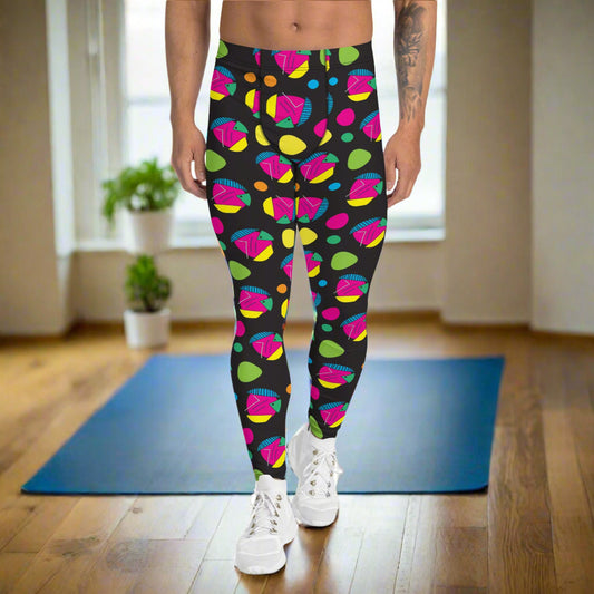 Man from the waist down wearing men's Funk Party funky festival yoga leggings standing facing forwards with his hands by his sides. The man is standing on a wooden floor in an apartment with light streaming in from a large window behind him. There is a blue gym or yoga mat and two small house plants near to the left of the window. The funk party pattern  is an abstract pattern in vibrant pink, orange, yellow, green and blue in a spotty type arrangement on a black background