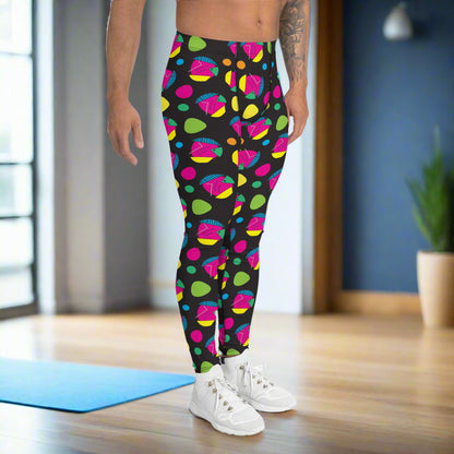 Man from the waist down wearing men's Funk Party funky festival yoga leggings standing facing to the right with his hands by his sides. The man is standing on a wooden floor in an apartment with light streaming in from a large window to the left of him. There is a blue gym or yoga mat behind him and a small house plant to the right against a blue wall. The funk party pattern  is an abstract pattern in vibrant pink, orange, yellow, green and blue in a spotty type arrangement on a black background