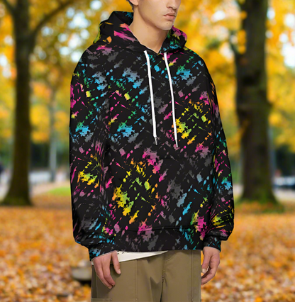 Man from the chin down to the waist, posing standing facing to the right wearing a 100% cotton vibrant Minstrel Magic tie-dye all-over print hoodie. He is standing outside amongst autumnal trees with golden leaves blanketing the ground. The hoodie is black with splashes of bright pink, orange yellow, green, blue and grey. It is a bold fun design for everyday wear.