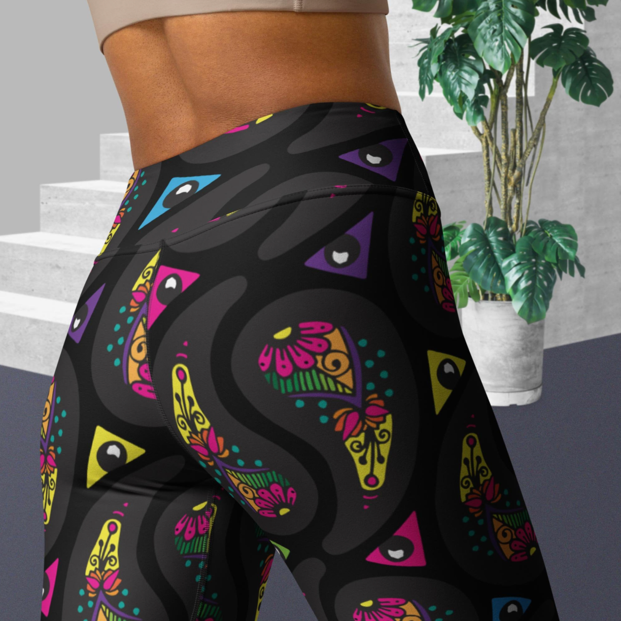 Close up of a woman from the upper back down to thighs, wearing high-waist Black Paisley Party funky festival yoga leggings standing facing away. The woman is standing on a grey floor in front of some white steps and a tall green plant to the right. The paisley party pattern is seen in more detail showing a colourful repeating large paisley pattern, made up of vibrant colours including pink, orange, yellow, green, blue and purple. In between the paisley print are colourful triangles in the same colours