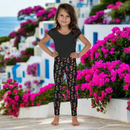 A young young girl wearing kids size black paisley party funky festival leggings, poses facing forwards. She is standing on a stone path that leads down to the sea. White and blue houses and colourful pink flowers create an idyllic scene.. The paisley party pattern  is a colourful repeating large paisley pattern, made up of vibrant colours including pink, orange, yellow, green, blue and purple. In between the paisley print are colourful triangles in the same colours