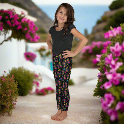 A young young girl wearing kids size black paisley party funky festival leggings, poses facing to the left. She is standing on a stone path that leads down the hill. Colourful pink flowers create an idyllic scene. The paisley party pattern  is a colourful repeating large paisley pattern, made up of vibrant colours including pink, orange, yellow, green, blue and purple. In between the paisley print are colourful triangles in the same colours