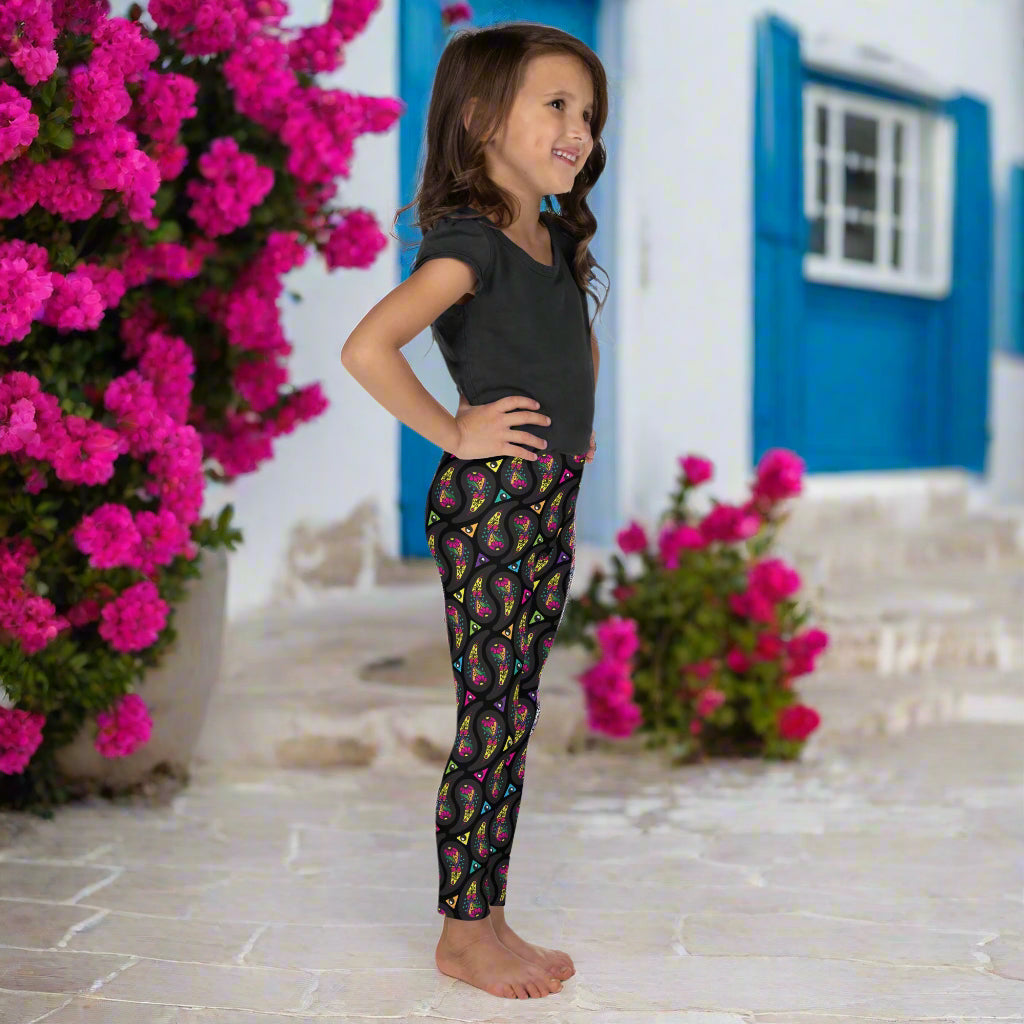 A young young girl wearing kids size black paisley party funky festival leggings, poses facing to the right. She is standing on a stone path outside some white and blue houses. Colourful pink flowers create an idyllic scene. The paisley party pattern  is a colourful repeating large paisley pattern, made up of vibrant colours including pink, orange, yellow, green, blue and purple. In between the paisley print are colourful triangles in the same colours