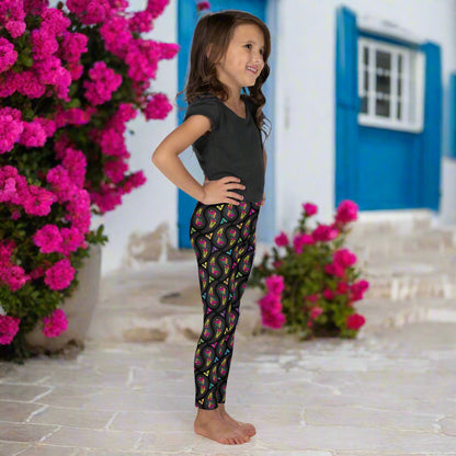 A young young girl wearing kids size black paisley party funky festival leggings, poses facing to the right. She is standing on a stone path outside some white and blue houses. Colourful pink flowers create an idyllic scene. The paisley party pattern  is a colourful repeating large paisley pattern, made up of vibrant colours including pink, orange, yellow, green, blue and purple. In between the paisley print are colourful triangles in the same colours