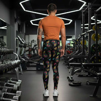 Man from the waist down wearing men's black paisley party funky festival yoga leggings standing facing away with his hands by his sides. He is wearing white trainers and is standing on a countryside green marked tennis court with trees and hedges in the background. The paisley party pattern is a colourful repeating large paisley pattern, made up of vibrant colours including pink, orange, yellow, green, blue and purple. In between the paisley print are colourful triangles in the same colours. 
