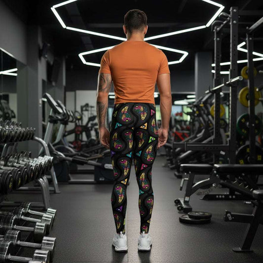 Man from the waist down wearing men's black paisley party funky festival yoga leggings standing facing away with his hands by his sides. He is wearing white trainers and is standing on a countryside green marked tennis court with trees and hedges in the background. The paisley party pattern is a colourful repeating large paisley pattern, made up of vibrant colours including pink, orange, yellow, green, blue and purple. In between the paisley print are colourful triangles in the same colours. 