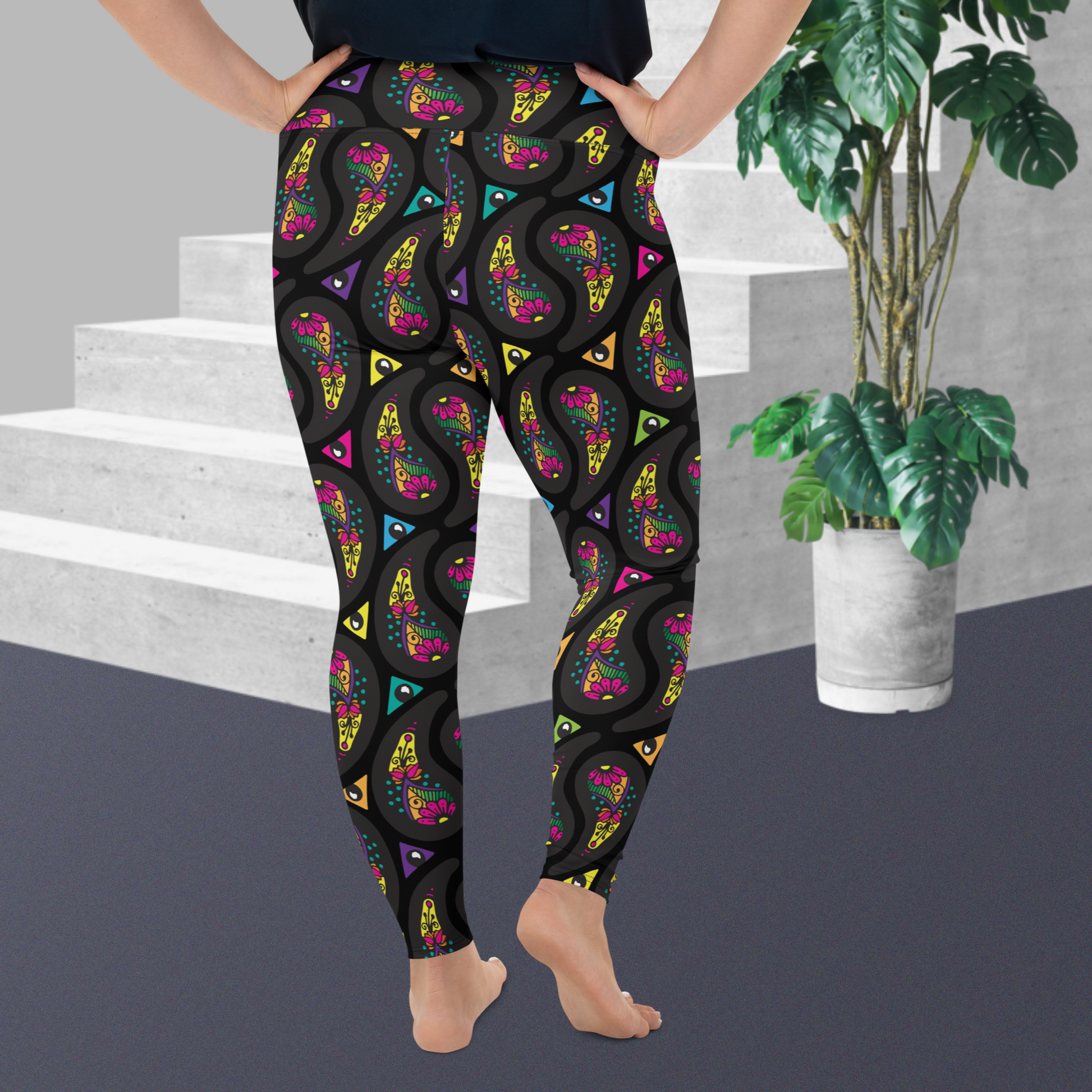 A curvy woman from the waist down wearing high-waist Black Paisley Party funky festival yoga leggings standing facing away with both hands on her hips. The woman is standing on a grey floor in front of some white steps with a tall green plant to the right. The paisley party pattern  is a colourful repeating large paisley pattern, made up of vibrant colours including pink, orange, yellow, green, blue and purple. In between the paisley print are colourful triangles in the same colours