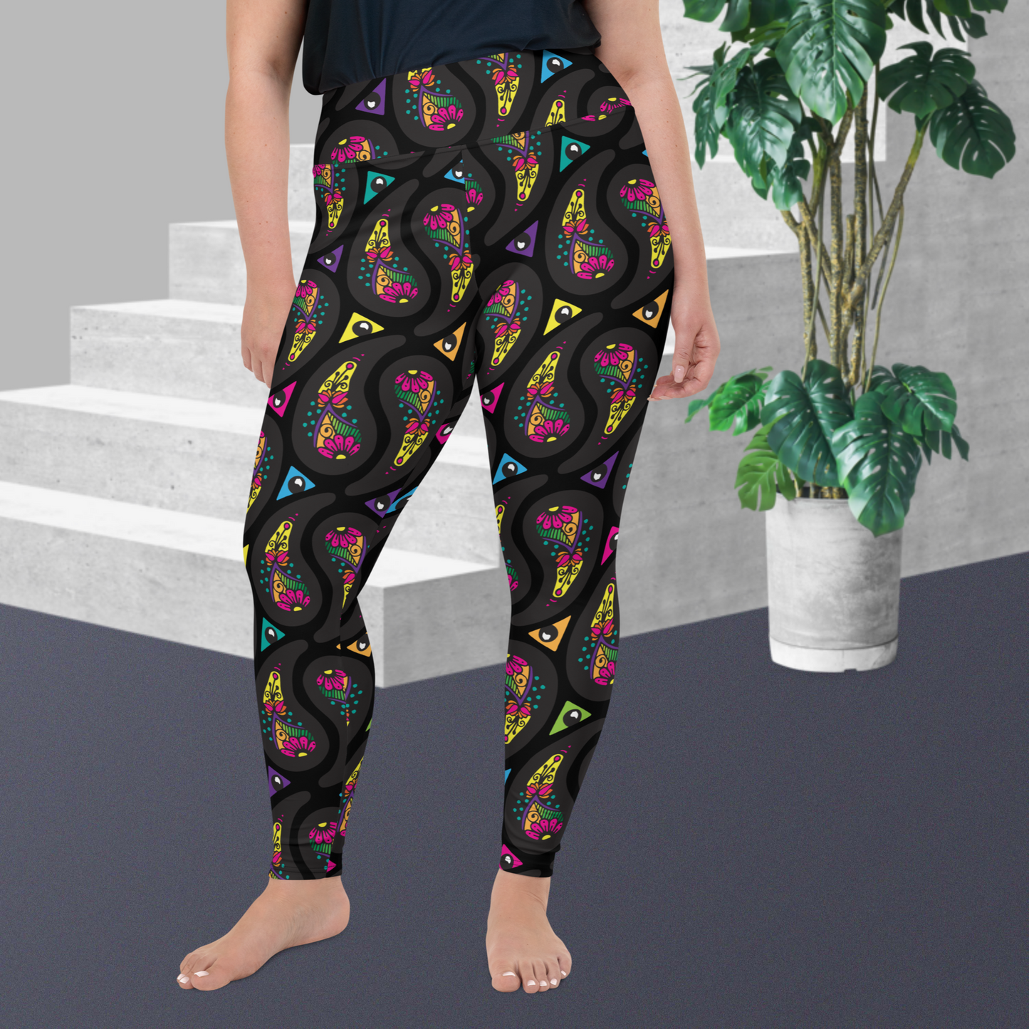 A curvy woman from the waist down wearing high-waist Black Paisley Party funky festival yoga leggings standing facing forwards with her hands by her sides. The woman is standing on a grey floor in front of some white steps with a tall green plant to the right. The paisley party pattern  is a colourful repeating large paisley pattern, made up of vibrant colours including pink, orange, yellow, green, blue and purple. In between the paisley print are colourful triangles in the same colours