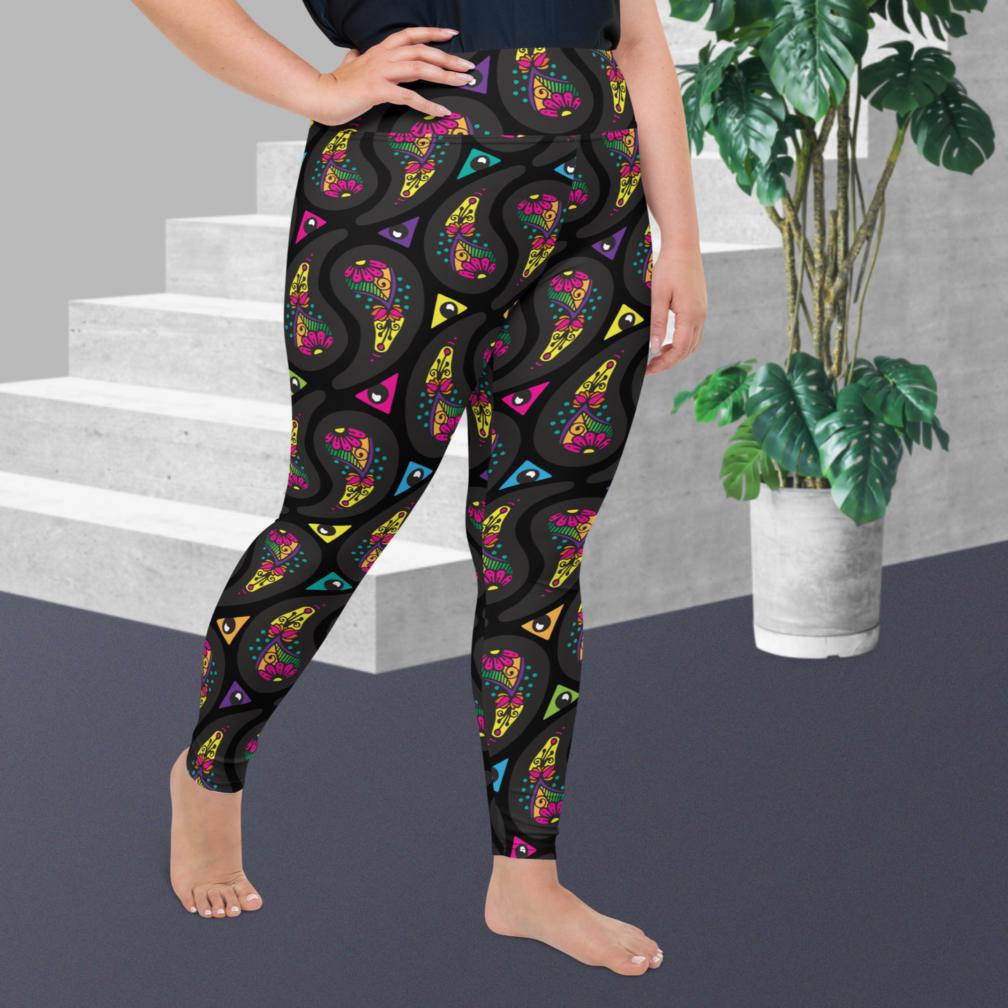A curvy woman from the waist down wearing high-waist Black Paisley Party funky festival yoga leggings standing facing right with one hand on her hip. The woman is standing on a grey floor in front of some white steps with a tall green plant to the right. The paisley party pattern  is a colourful repeating large paisley pattern, made up of vibrant colours including pink, orange, yellow, green, blue and purple. In between the paisley print are colourful triangles in the same colours