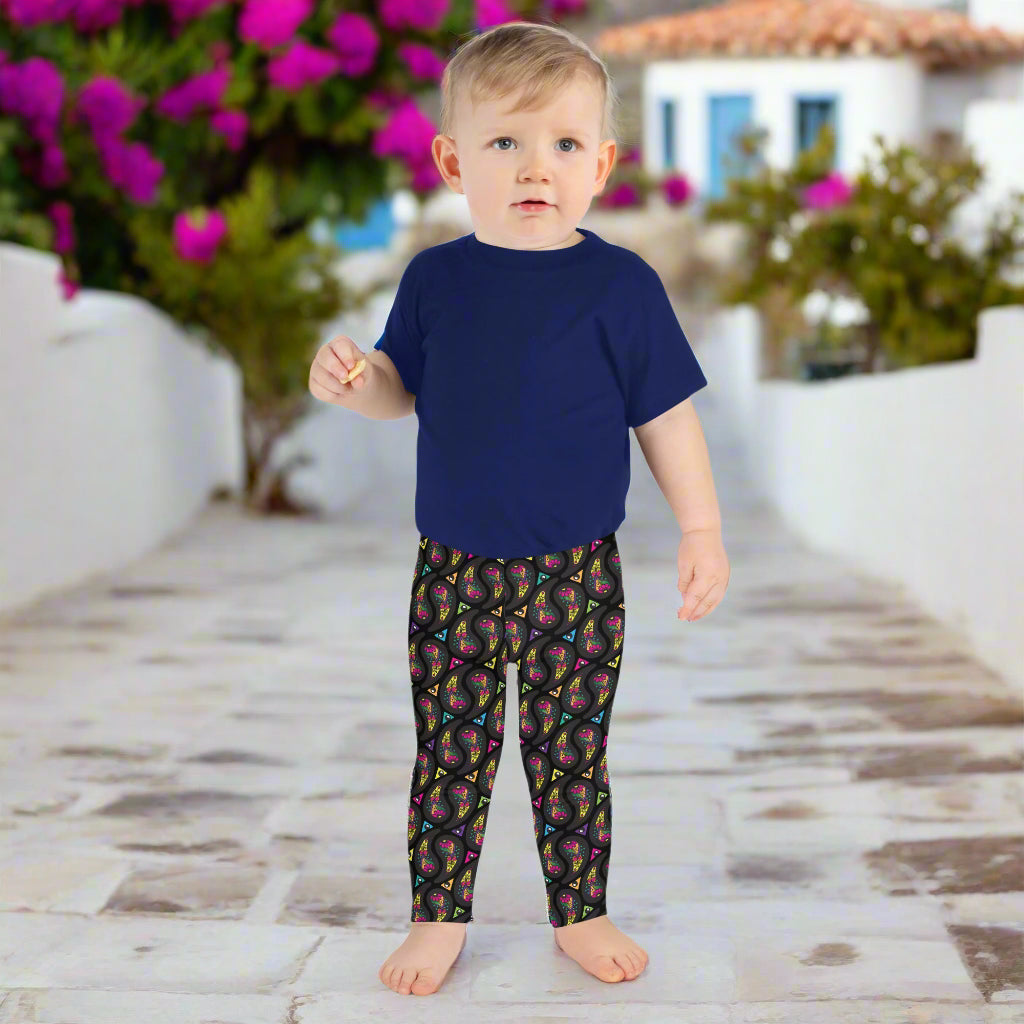 A toddler wearing kids size black paisley party funky festival leggings, poses facing forwards. He is standing on a stone path that leads to a white and blue house with bright pink flowers growing in the background. The paisley party pattern  is a colourful repeating large paisley pattern, made up of vibrant colours including pink, orange, yellow, green, blue and purple. In between the paisley print are colourful triangles in the same colours