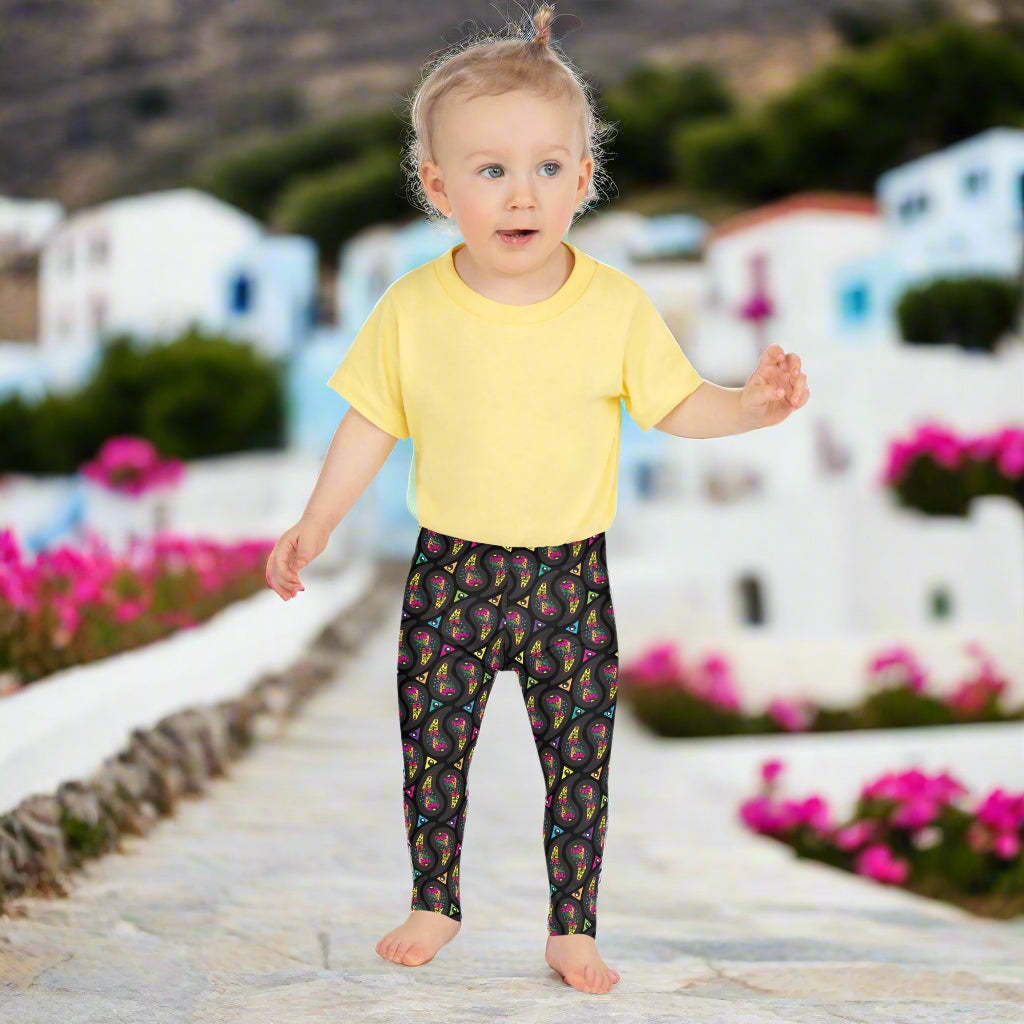 A toddler wearing kids size black paisley party funky festival leggings, poses facing forwards. They are standing on a stone path that leads away from some white and blue houses and colourful pink flowers are scattered around in the background. The paisley party pattern  is a colourful repeating large paisley pattern, made up of vibrant colours including pink, orange, yellow, green, blue and purple. In between the paisley print are colourful triangles in the same colours
