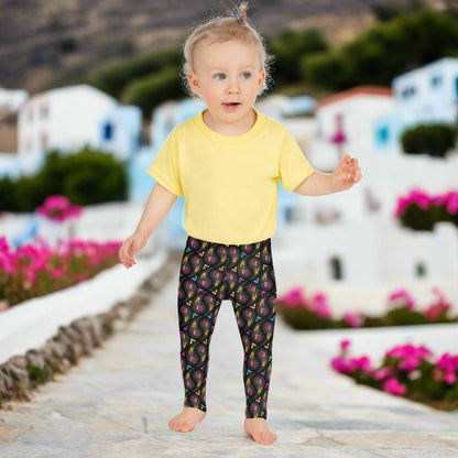 A toddler wearing kids size black paisley party funky festival leggings, poses facing forwards. They are standing on a stone path that leads away from some white and blue houses and colourful pink flowers are scattered around in the background. The paisley party pattern  is a colourful repeating large paisley pattern, made up of vibrant colours including pink, orange, yellow, green, blue and purple. In between the paisley print are colourful triangles in the same colours