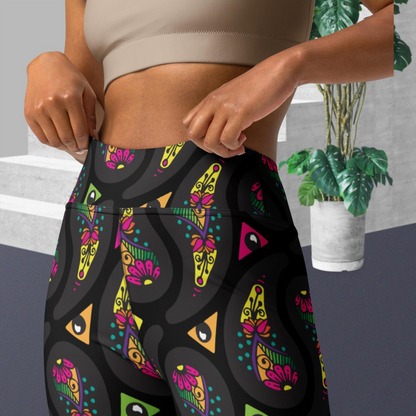Close up of a woman from the chest down to thighs, wearing high-waist Black Paisley Party funky festival yoga leggings standing facing forward. The woman is standing on a grey floor in front of some white steps and a tall green plant to the right. The paisley party pattern is seen in more detail showing a colourful repeating large paisley pattern, made up of vibrant colours including pink, orange, yellow, green, blue and purple. In between the paisley print are colourful triangles in the same colours