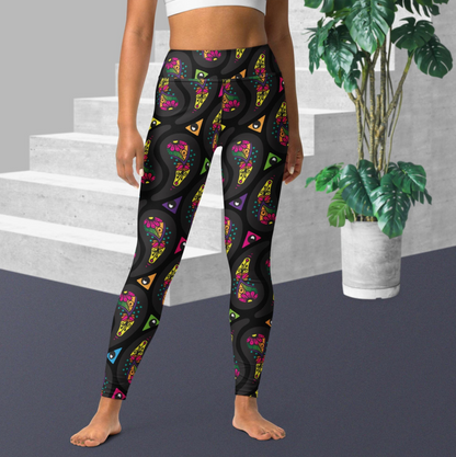 Woman from the waist down wearing high-waist Black Paisley Party funky festival yoga leggings standing facing forwards with her hands by her sides. The woman is standing on a grey floor in front of some white steps with a tall green plant to the right. The paisley party pattern  is a colourful repeating large paisley pattern, made up of vibrant colours including pink, orange, yellow, green, blue and purple. In between the paisley print are colourful triangles in the same colours
