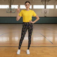 A young person from the waist down wearing youth size black paisley party funky festival leggings, poses facing away. They are standing on a stone path that leads down to the sea. White and blue houses and colourful pink , orange and blue flowers create an idyllic scene.. The paisley party pattern  is a colourful repeating large paisley pattern, made up of vibrant colours including pink, orange, yellow, green, blue and purple. In between the paisley print are colourful triangles in the same colours