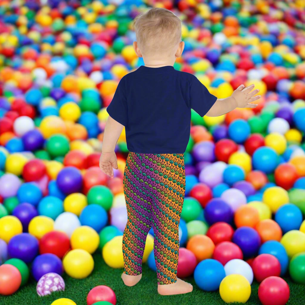 A toddler wearing kids size fire tribal frog funky festival leggings and a navy blue shirt stands facing away looking at a ball pit filled with multicoloured plastic balls. The fire tribal frog pattern  is an abstract pattern with a blended orange, pink and purple background.