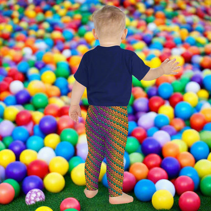 A toddler wearing kids size fire tribal frog funky festival leggings and a navy blue shirt stands facing away looking at a ball pit filled with multicoloured plastic balls. The fire tribal frog pattern  is an abstract pattern with a blended orange, pink and purple background.