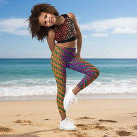 A young girl wearing kids size fire tribal frog funky festival leggings, poses on one leg facing forwards. She is smiling and standing in front of a background of pastel coloured lights that look like a galaxy. The fire tribal frog pattern  is an abstract pattern with a blended orange, pink and purple background.