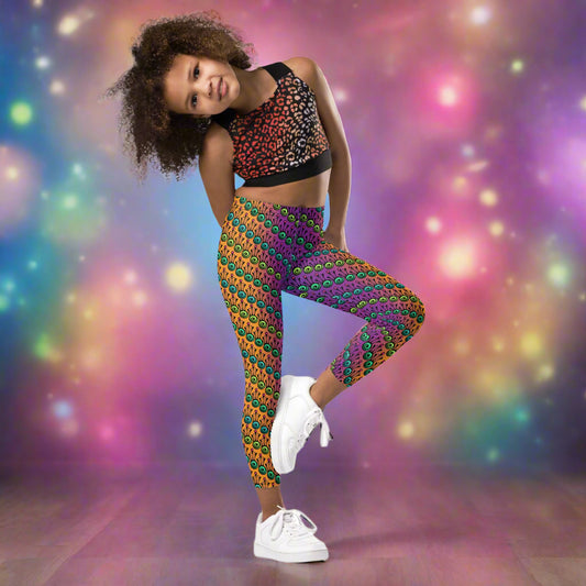 A young girl wearing kids size fire tribal frog funky festival leggings, poses on one leg facing forwards. She is smiling and standing in front of a background of pastel coloured lights that look like a galaxy. The fire tribal frog pattern  is an abstract pattern with a blended orange, pink and purple background.