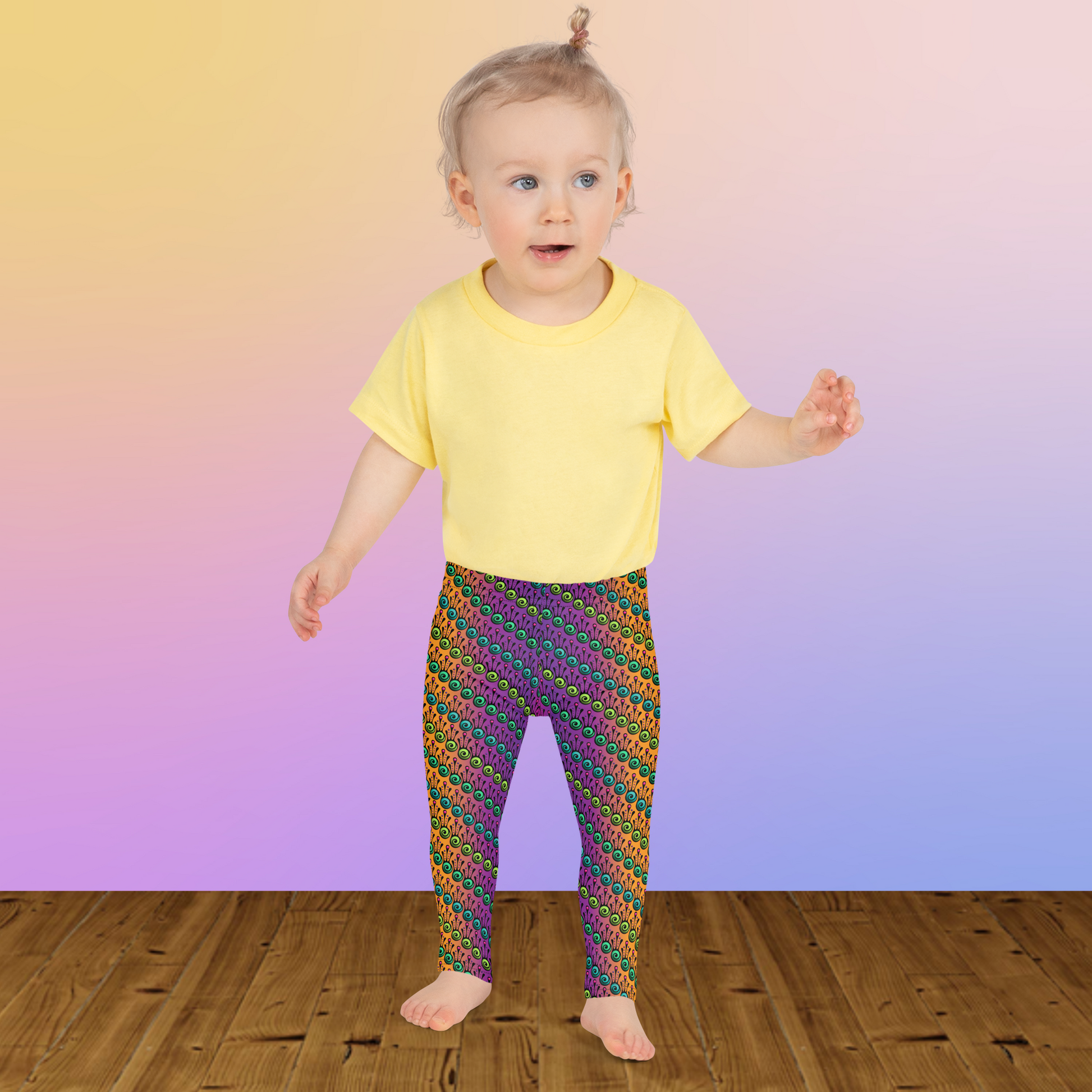 A toddler wearing kids size fire tribal frog funky festival leggings and a pale yellow t-shirt stands on a wooden floor facing forwards. She is standing in front of a pale pastel pink, purple and yellow coloured background. The fire tribal frog pattern  is an abstract pattern with a blended orange, pink and purple background.